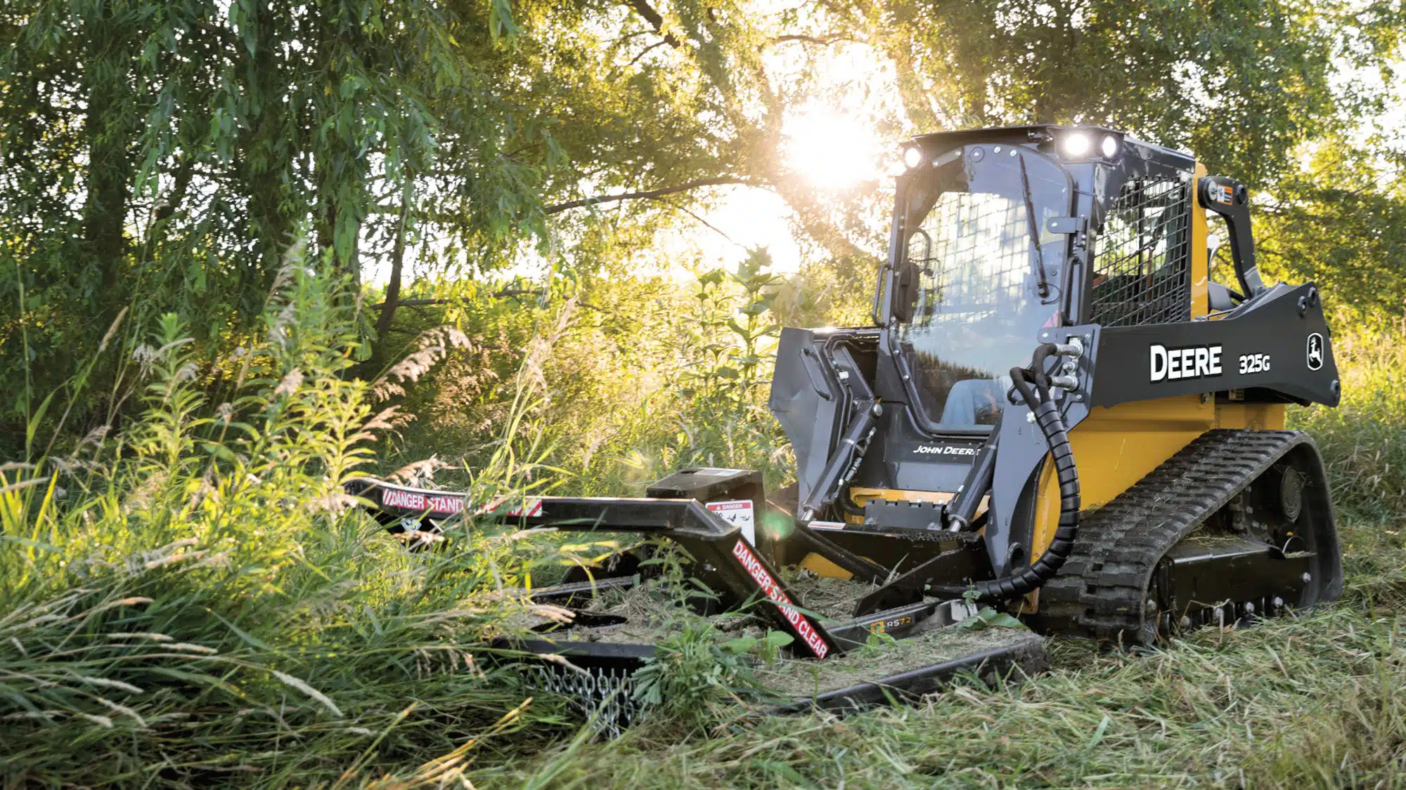 John Deere 325G compact track loader with a rotary brush cutter clearing brush in a grassy field